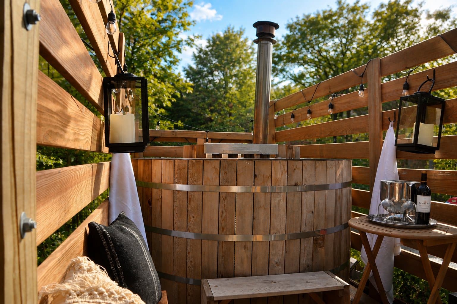 Cedar hot tub in daylight surrounded by trees, with a lantern and wine service nearby