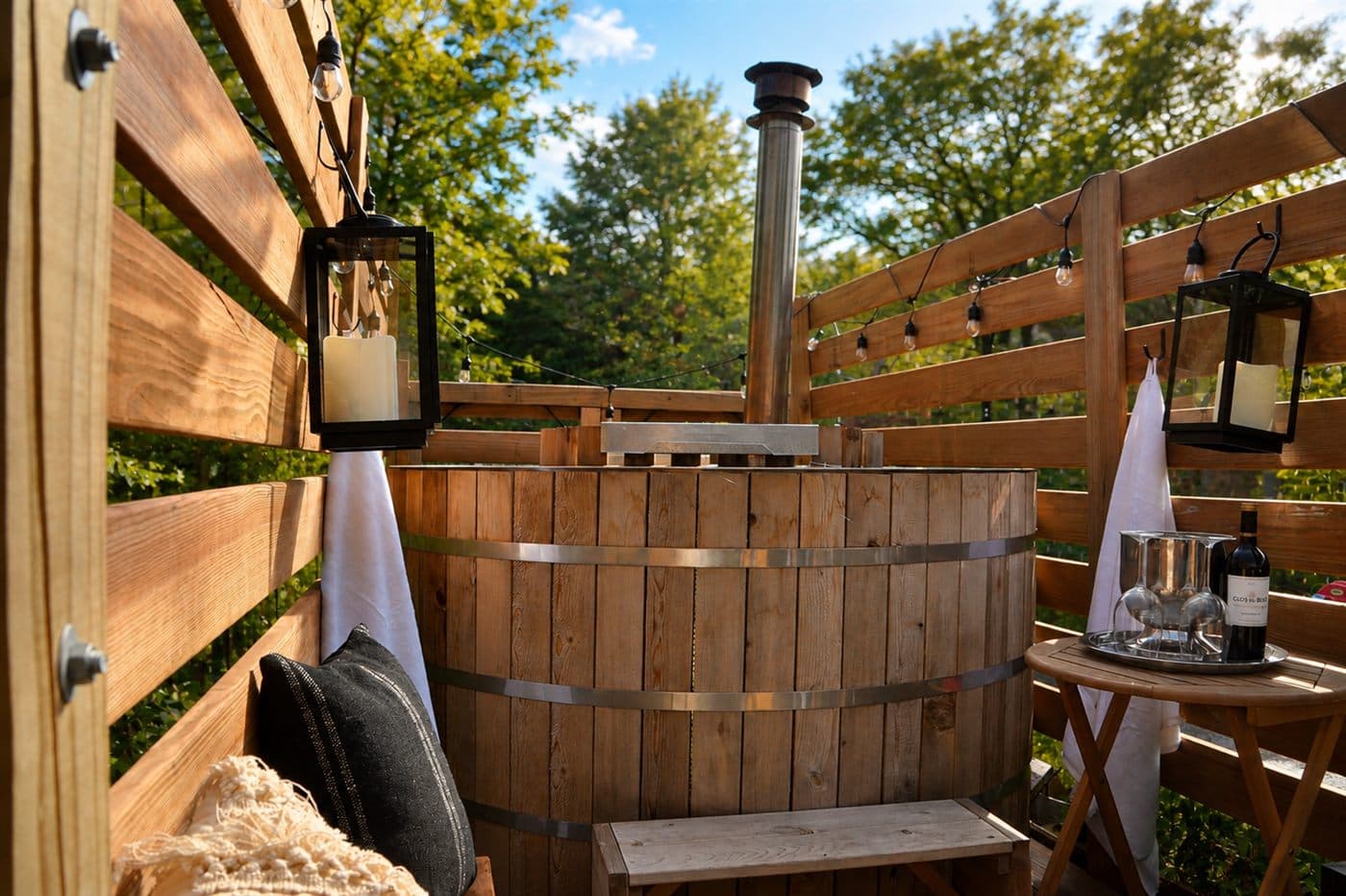 Cedar hot tub in daylight surrounded by trees, with a lantern and wine service nearby